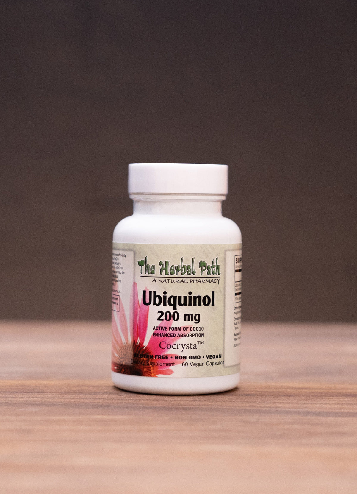 A white bottle of The Medicine Patch GeEnzyme Q10 Softgel 300 mg stands centered on a wooden tabletop against a dark background, with blurred herbal props on both sides.