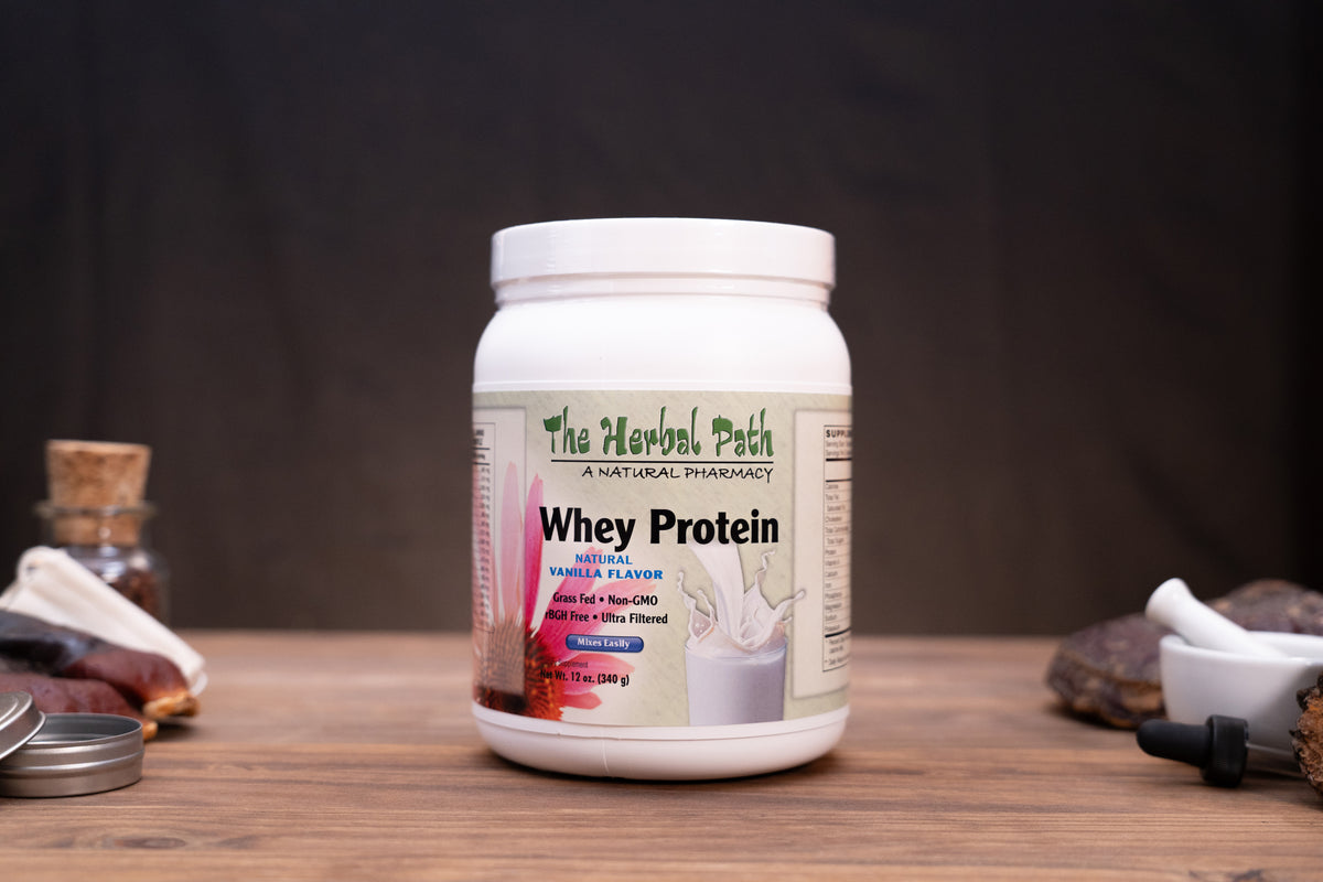 A white plastic tub of 'The Herbal Path' Whey Protein powder sits on a wooden table. The label indicates it is Vanilla Flavored and contains 2 lbs of protein powder.