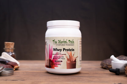 A white tub of The Herbal Path Whey Protein powder on a wooden table, surrounded by small natural ingredients and kitchen props against a dark background.