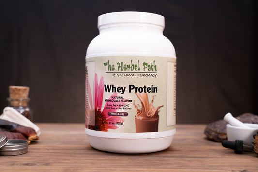 A white tub of Whey Protein by The Herbal Path on a wooden surface with a dark background, flanked by small props like a cork stopper, a small bottle, and a dropper.