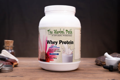 A white plastic tub of 'The Herbal Path' Whey Protein in Vanilla Flavored, displayed on a wooden surface with natural elements like a mortar and pestle, dried flowers, and a small bottle in the background.