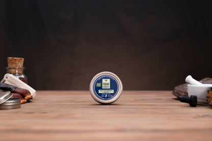 A small round tin of beard balm stands upright on a wooden tabletop, centered against a dark brown background. The tin has a blue and white label with the brand name 'Beard Care' visible.