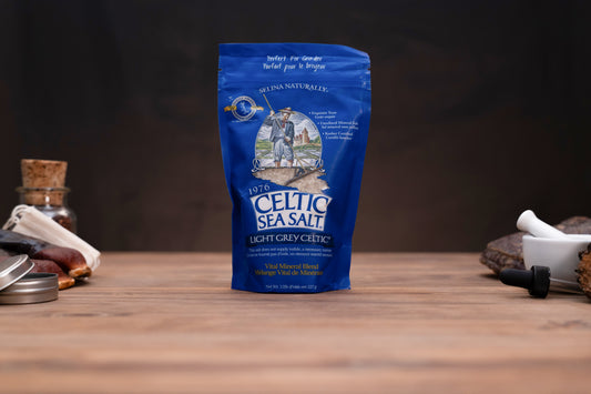 A blue stand-up pouch of Celtic Sea Salt stands on a wooden surface against a dark background, with small kitchen props visible on both sides.