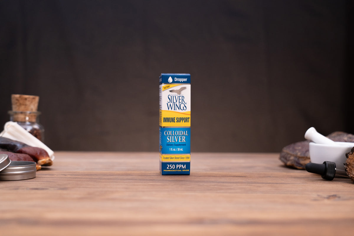 A small box of Dropper Silver Wings Bone Support Collagen Silver centered on a wooden tabletop, with a dark blurred background and rustic props on both sides.