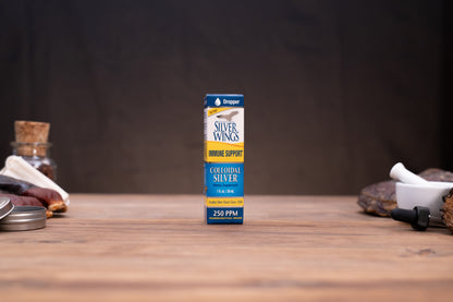A small box of Dropper Silver Wings Bone Support Collagen Silver centered on a wooden tabletop, with a dark blurred background and rustic props on both sides.