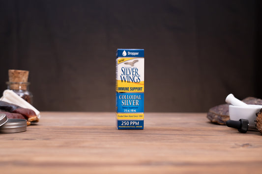 A box of Draper Silver Wings Colloidal Silver stands centered on a wooden tabletop against a dark backdrop, with small rustic props visible on both sides.