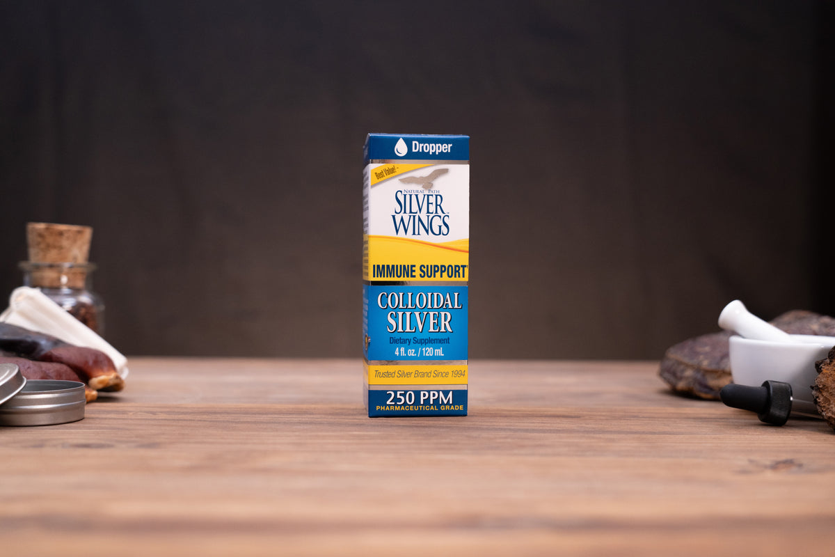 A box of Silver Wings Immune Support Colloidal Silver stands centered on a wooden tabletop against a dark backdrop, with small blurred props visible on both sides.