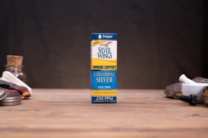 A box of Silver Wings Immune Support Colloidal Silver stands centered on a wooden tabletop against a dark backdrop, with small blurred props visible on both sides.