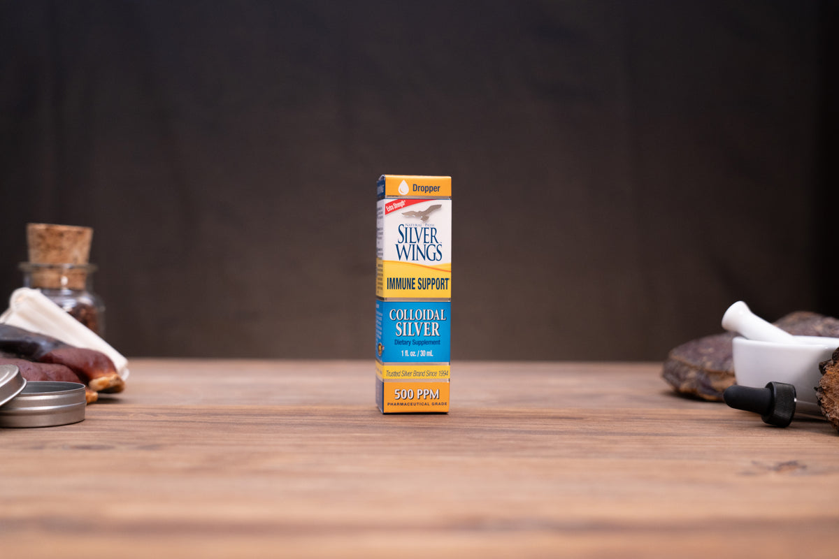 A small box of Silver Wings Colourful Silver immune support capsules stands centered on a wooden tabletop against a dark backdrop, with blurred jars and a white mortar-like object on either side.