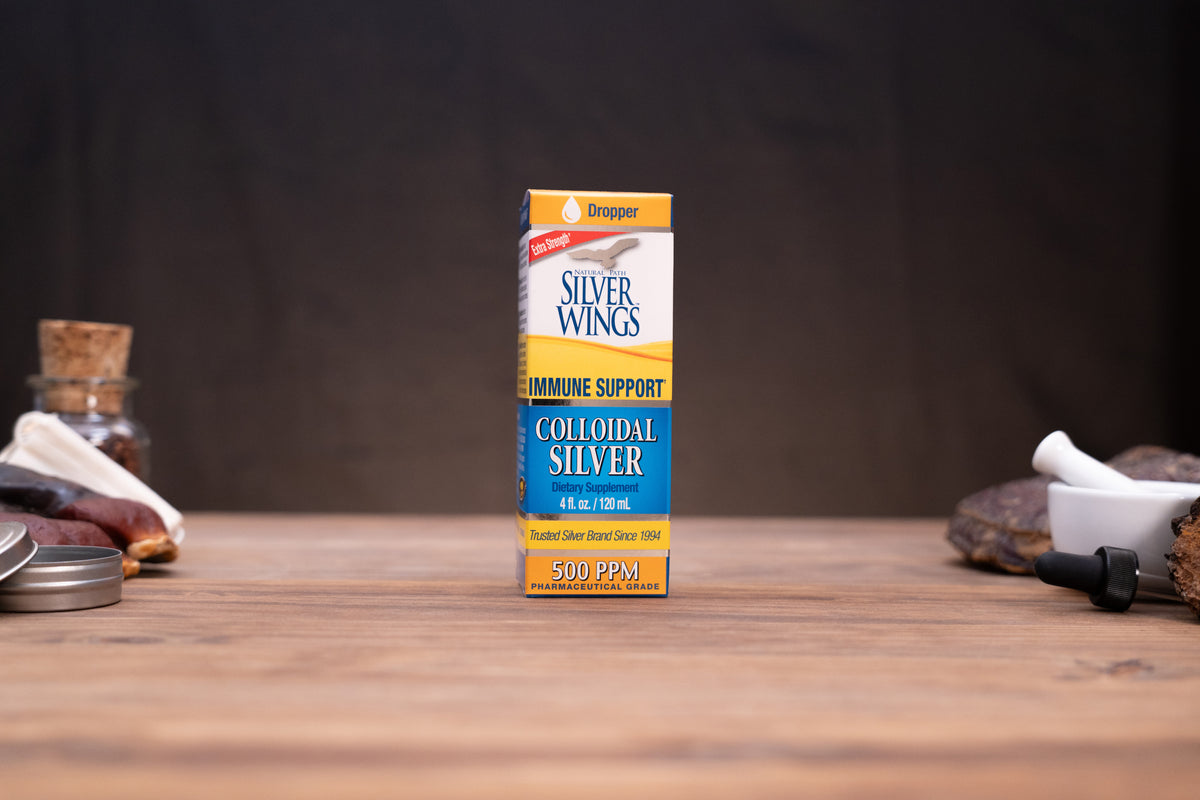 Silver Wings Immune Support Colloidal Silver 500 PPM dropper bottle standing on a wooden surface with rustic herbal props blurred in the background.