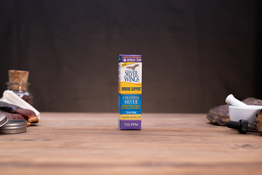 A box of Silver Wings Whole Support Calmoseptic Silver 150 PPM spray top, standing upright on a wooden tabletop with blurred rustic props on either side against a dark background.