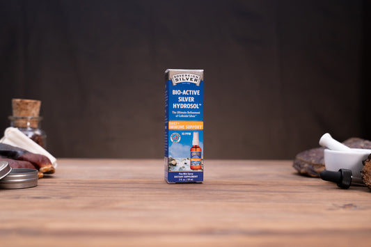 A box of Silver Silver Bio-Active Silver Hydrosol standing upright on a wooden tabletop, with blurred rustic props on both sides against a dark backdrop.