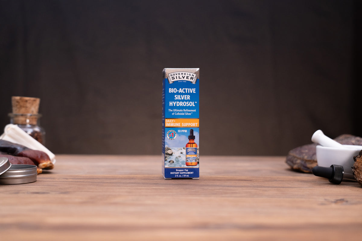 A box of Silver Silver Bio-Active Silver Hydrogel standing upright on a wooden tabletop, with blurred natural props on both sides against a dark backdrop.