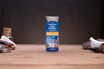 A box of Love Action Silver Bio-Active Silver Hydrogel for skin support, standing upright on a wooden tabletop with a dark, moody background and small blurred props on either side.