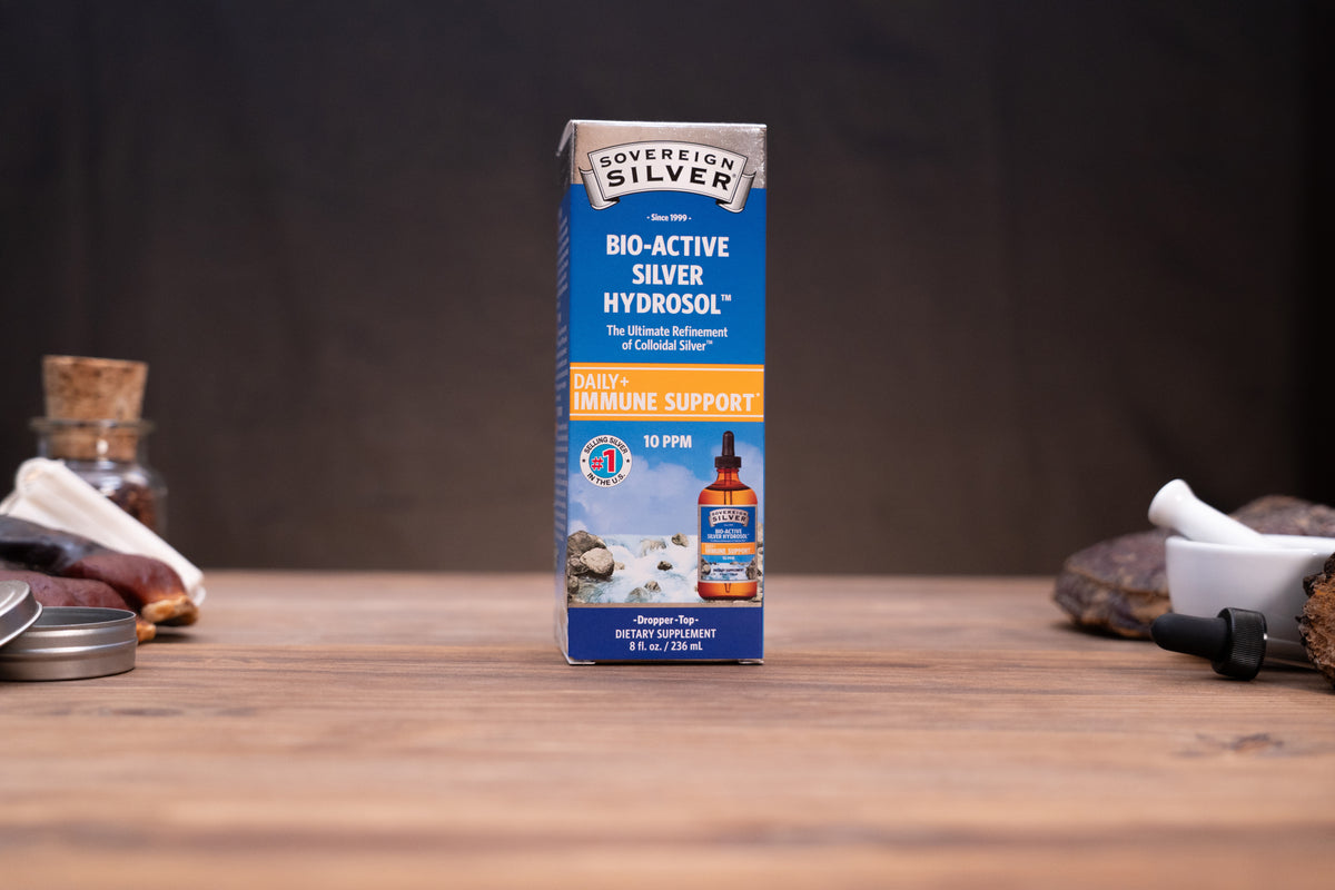A box of Sovereign Silver Bio-Active Silver Hydrocolol dietary supplement stands on a wooden tabletop against a dark, blurred background. The packaging is primarily blue and white with orange accents, and a small bottle is pictured on the front.