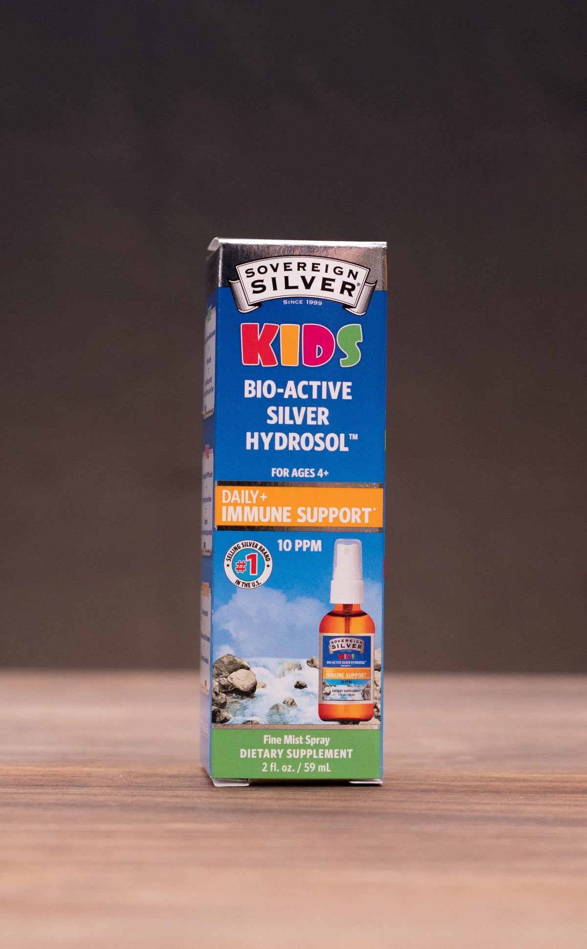 A box of Silver Kids Big-Active Silver Hydrators standing upright on a wooden tabletop, with a dark draped backdrop behind it and small rustic props blurred at the sides.