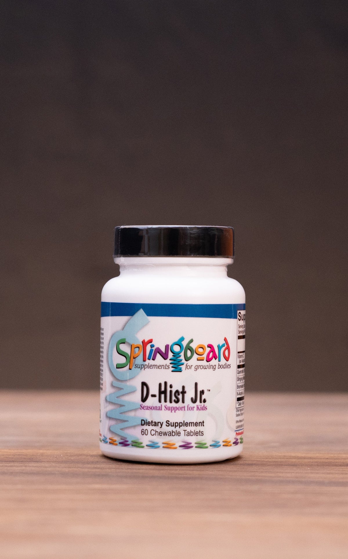 A white bottle of Springbos D-Hut Jc dietary supplement with a black cap, centered on a wooden surface with a dark background and blurred herbal props.