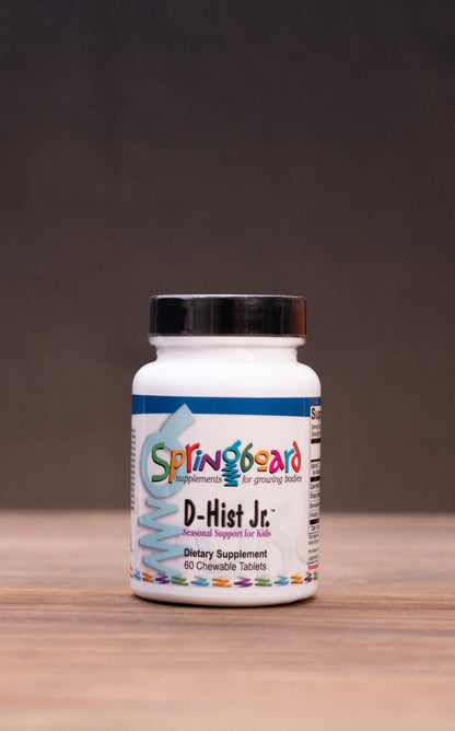 A white bottle of Springbos D-Hut Jc dietary supplement with a black cap, centered on a wooden surface with a dark background and blurred herbal props.