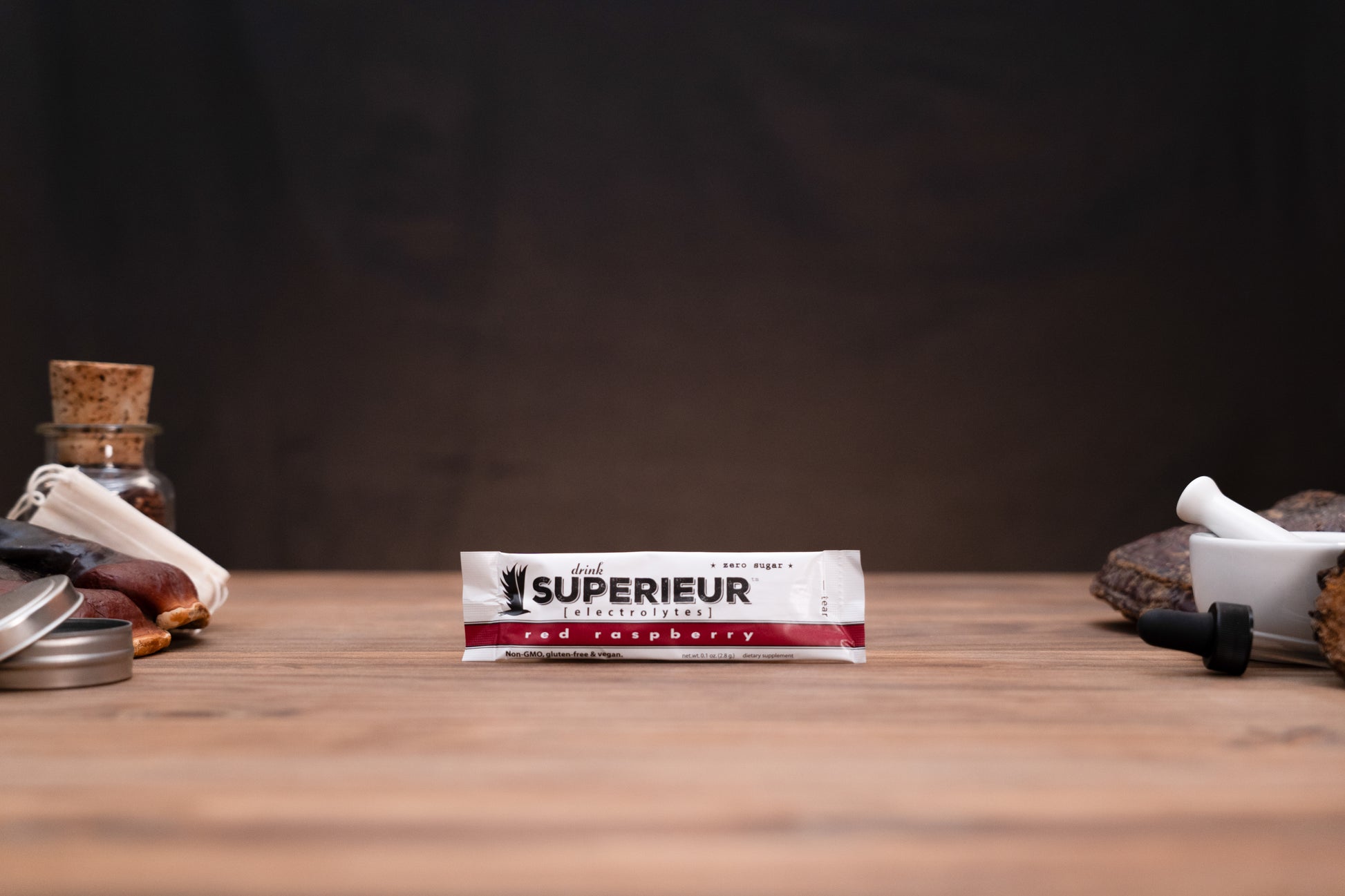 A package of 'SUPERIEUR' red raspberry ice cream stands centered on a wooden table, flanked by blurred rustic props including a glass bottle with a cork stopper, metal tins, and a white mortar and pestle against a dark background.