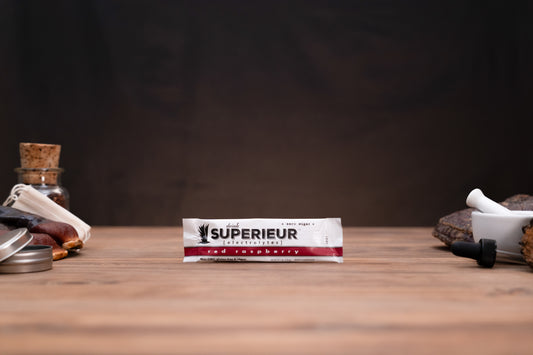 A package of 'SUPERIEUR' red raspberry ice cream stands centered on a wooden table, flanked by blurred rustic props including a glass bottle with a cork stopper, metal tins, and a white mortar and pestle against a dark background.