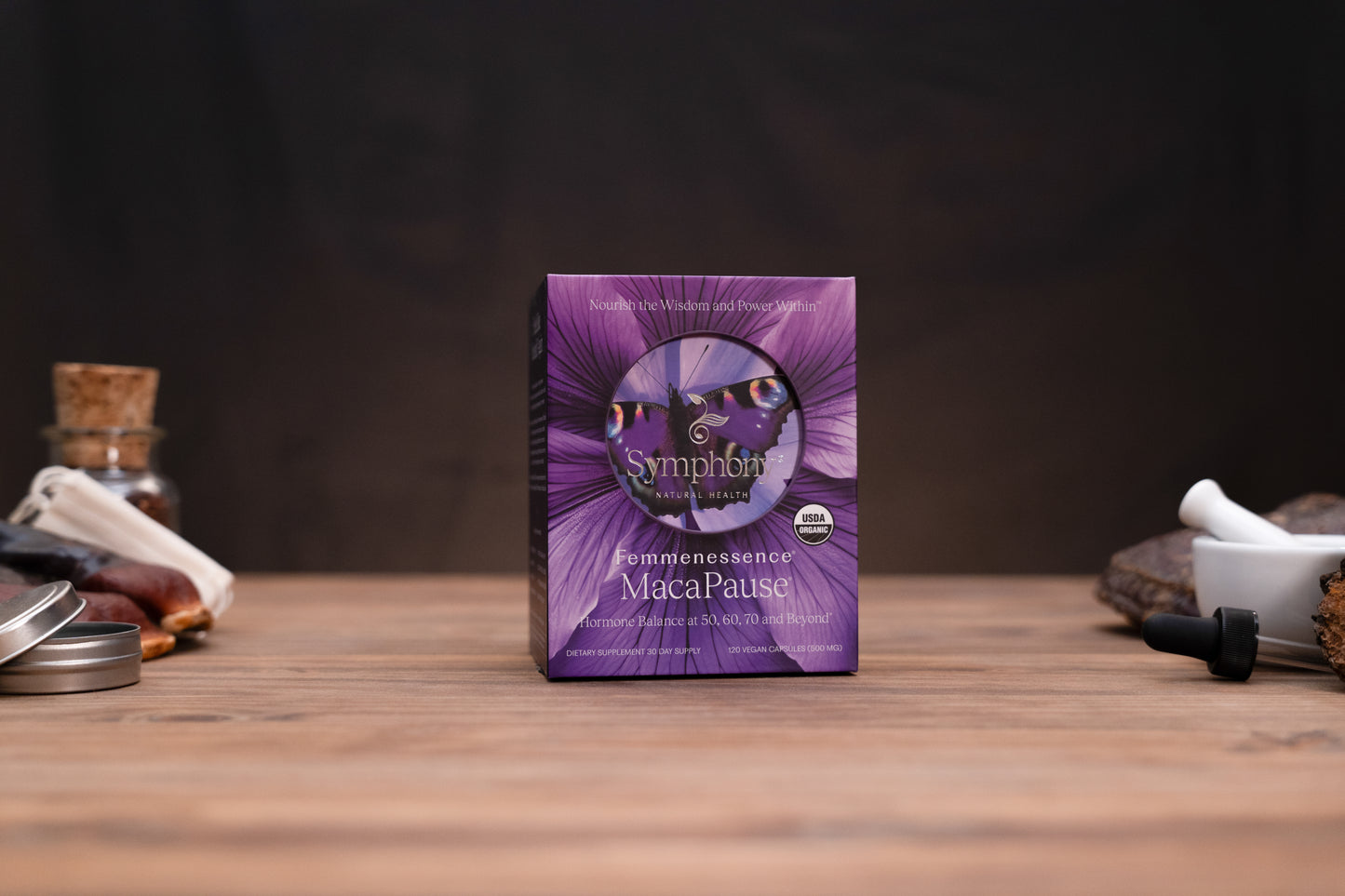 Symphonics Maca Pause supplement box in purple packaging, standing on a wooden surface with blurred herbal props in the background.