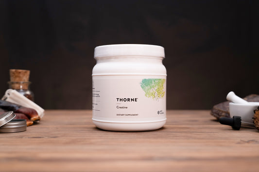 A white plastic jar of Thorne Creatine dietary supplement sits on a rustic wooden table, surrounded by subtle herbal and laboratory props against a dark, moody background.