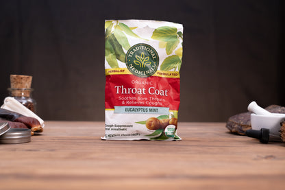 A package of Traditional Medicines Organic Throat Coat Eucalyptus Mint herbal syrup stands on a wooden surface against a dark background, with small herbal-themed props visible on both sides.