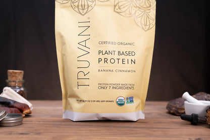 A stand-up pouch of Truvani Certified Organic Plant Based Protein in Banana Cinnamon flavor, displayed on a wooden surface with small kitchen props like a mortar and pestle, spices, and a jar in the background.