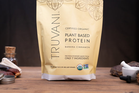 A stand-up pouch of Truvani Certified Organic Plant Based Protein in Banana Cinnamon flavor, displayed on a wooden surface with small kitchen props like a mortar and pestle, spices, and a jar in the background.