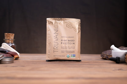 A bag of Truvani Plant Based Protein Chocolate Peanut Butter is centered on a wooden table, flanked by rustic props including a small glass bottle, a metal lid, and a white mortar and pestle against a dark background.