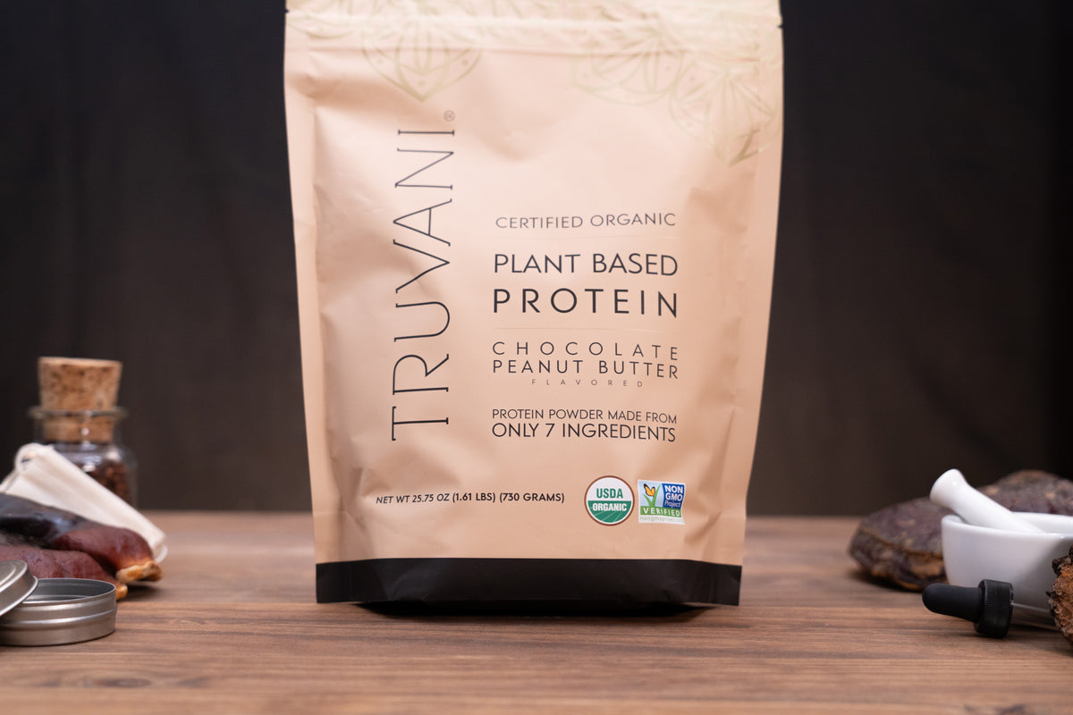 A stand-up pouch of Truvani Certified Organic Plant Based Protein in Chocolate Peanut Butter flavor, displayed on a wooden surface with small kitchen ingredients and a mortar and pestle in the background.