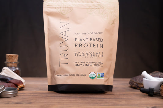 A stand-up pouch of Truvani Certified Organic Plant Based Protein in Chocolate Peanut Butter flavor, displayed on a wooden surface with small kitchen ingredients and a mortar and pestle in the background.