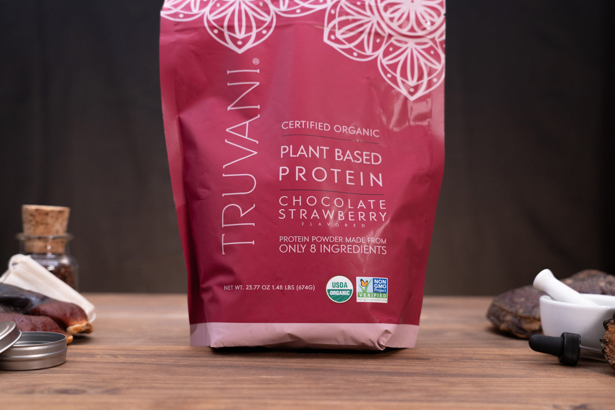A bag of Truvani Certified Organic Plant Based Protein in Chocolate Strawberry flavor, standing on a wooden surface with small ingredients and a mortar and pestle blurred in the background.