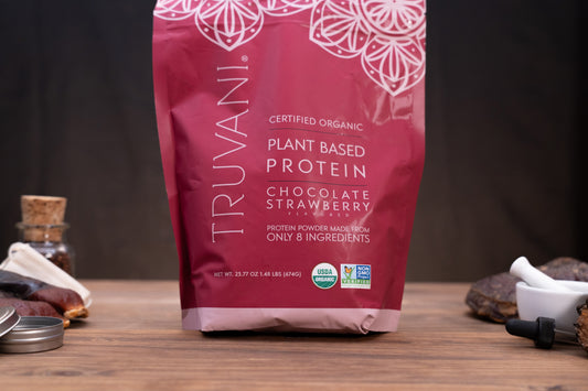 A bag of Truvani Certified Organic Plant Based Protein in Chocolate Strawberry flavor, standing on a wooden surface with small ingredients and a mortar and pestle blurred in the background.