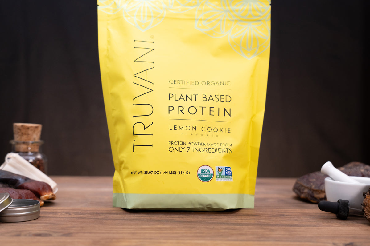 A yellow stand-up pouch of Truvani Plant Based Protein in Lemon Cookie flavor, standing on a wooden surface with small kitchen props like a dropper bottle, jar, and dried fruit around it.