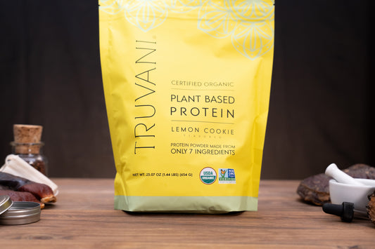 A yellow stand-up pouch of Truvani Plant Based Protein in Lemon Cookie flavor, standing on a wooden surface with small kitchen props like a dropper bottle, jar, and dried fruit around it.