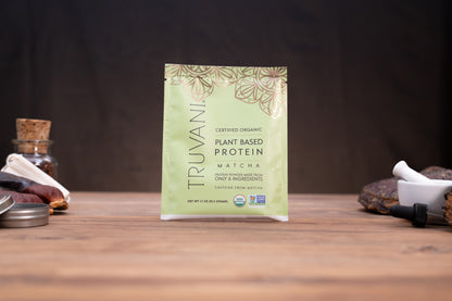 A single packet of Truvani Certified Organic Plant Based Protein Matcha is centered on a wooden surface with a dark, moody background and small rustic props on either side.
