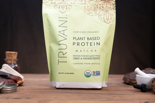 A 22 oz bag of Truvani Certified Organic Plant Based Protein Matcha, standing on a wooden surface with small kitchen props like a small bottle, cloth, and mortar and pestle in the background.