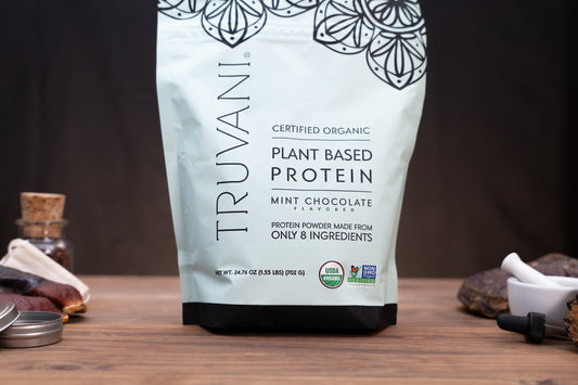A stand-up pouch of Truvani Certified Organic Plant Based Protein in Mint Chocolate Flavour, displayed on a wooden surface with small kitchen props like a mortar and pestle and dried fruit in the background.