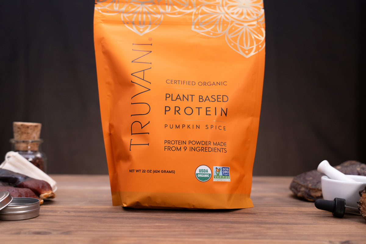 A stand-up pouch of Truvani Plant Based Protein in Pumpkin Spice flavor, displayed on a wooden surface with small kitchen props like a glass bottle, spoon, and dried ingredients in the background.
