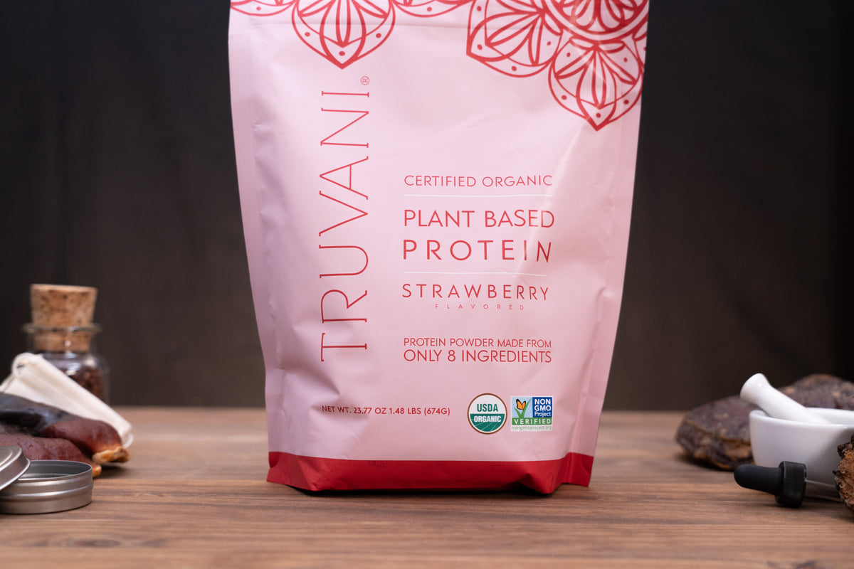 A stand-up pouch of Truvani Plant Based Protein Strawberry flavor, shown on a wooden surface with small kitchen or supplement props blurred in the background.