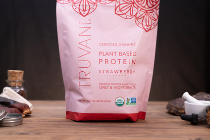 A stand-up pouch of Truvani Plant Based Protein Strawberry flavor, shown on a wooden surface with small kitchen or supplement props blurred in the background.