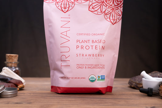 A stand-up pouch of Truvani Plant Based Protein Strawberry flavor, shown on a wooden surface with small kitchen or supplement props blurred in the background.