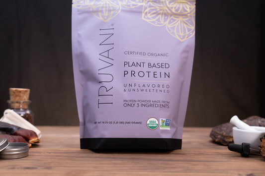 A bag of Truvani Certified Organic Plant Based Protein powder, unflavored and unsweetened, standing on a wooden surface with small kitchen props like a mortar and pestle and spices in the background.