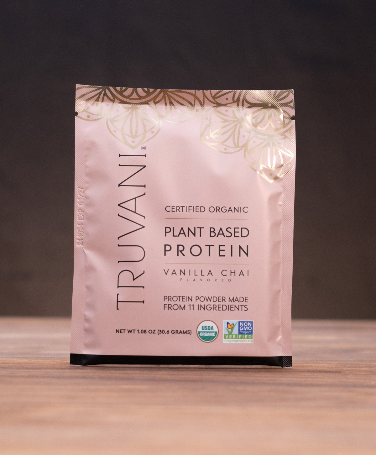 A stand-up pouch of Truvani Plant Based Protein Vanilla Chai stands on a wooden surface against a dark background, with small kitchen props visible on both sides.