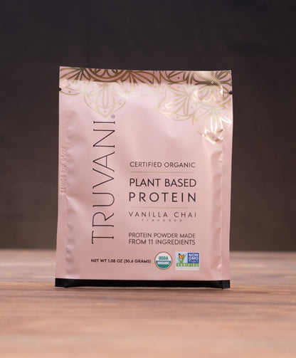 A stand-up pouch of Truvani Plant Based Protein Vanilla Chai stands on a wooden surface against a dark background, with small kitchen props visible on both sides.