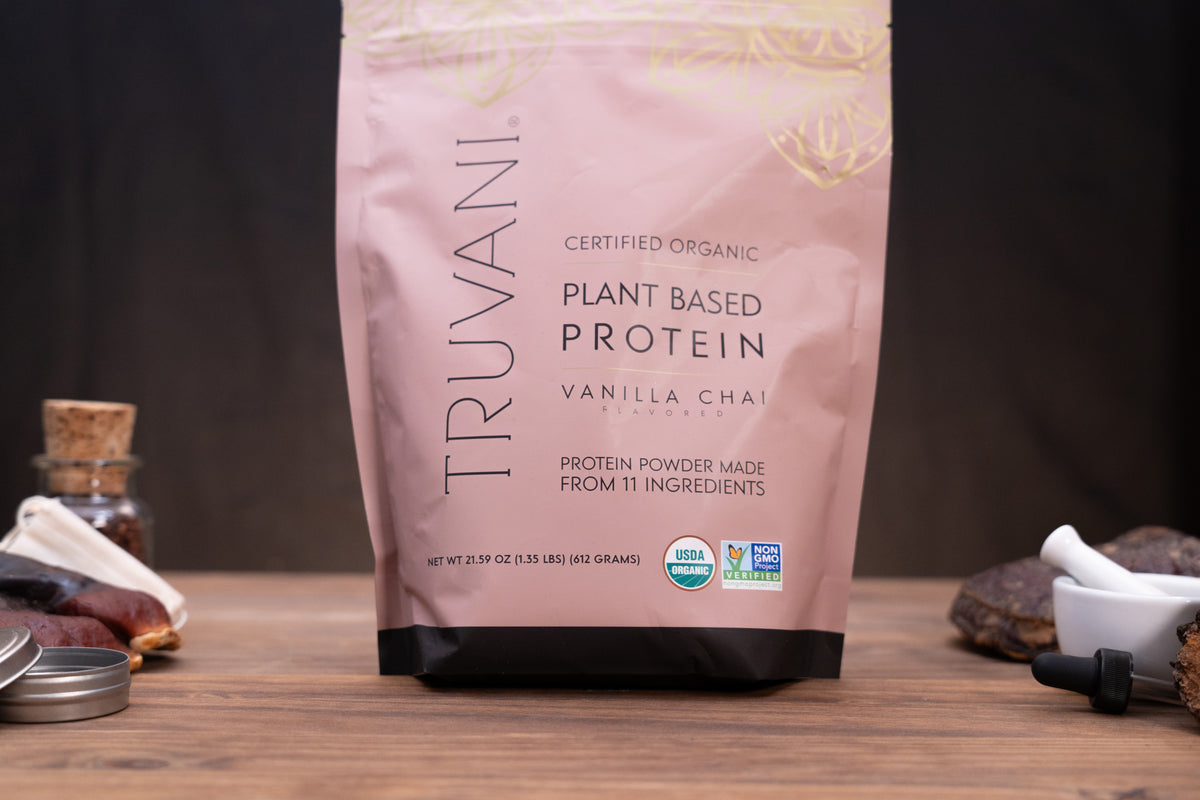 A pink stand-up pouch of Truvani Certified Organic Plant Based Protein Vanilla Chai flavor, standing on a wooden surface with small kitchen props like a mortar and pestle, dried fruit, and a small bottle in the background.