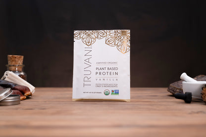 A package of Truvani Certified Organic Plant Based Protein Vanilla flavor, standing upright on a wooden table with rustic props like a cork stopper and mortar and pestle in the background.