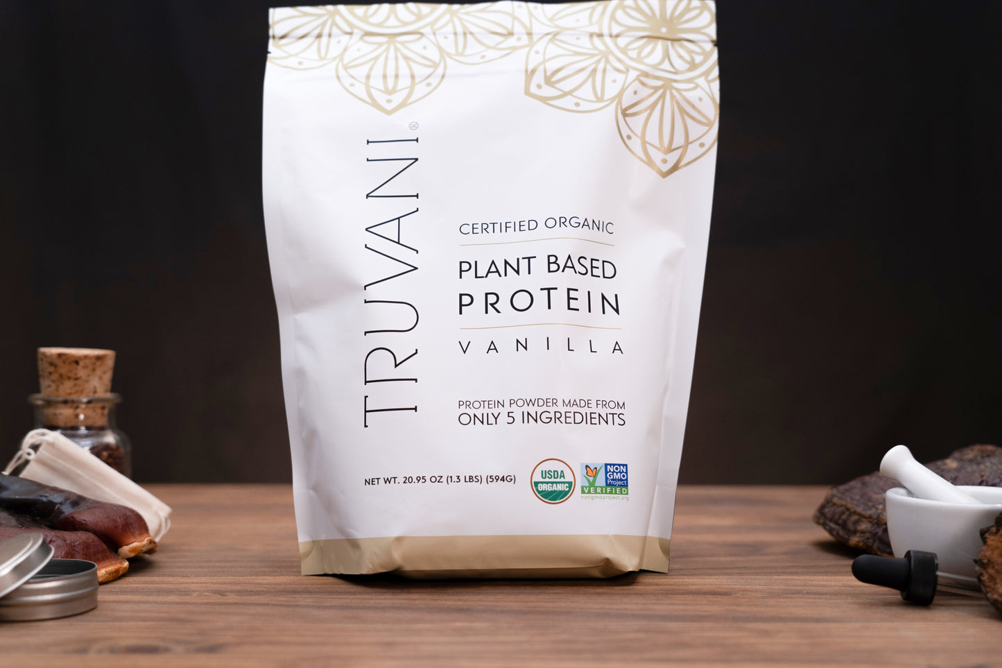 A bag of Truvani Plant Based Protein Vanilla powder stands on a wooden surface against a dark background, with small kitchen props like a bottle, spoon, and mortar and pestle partially visible at the sides.