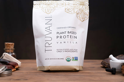 A bag of Truvani Plant Based Protein Vanilla powder stands on a wooden surface against a dark background, with small kitchen props like a bottle, spoon, and mortar and pestle partially visible at the sides.